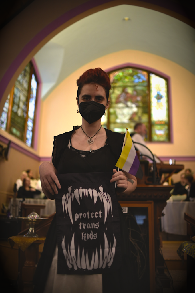 A nonbinary person with red hair and a black dress holding a black flag with white fangs and white text that reads Protect Trans Kids